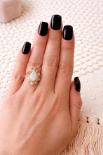 Load image into Gallery viewer, Hand with black nail polish wearing a gold ring with a opal stone on a textured surface.