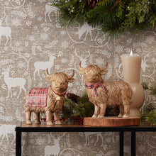 Load image into Gallery viewer, Two Highland Cow figurines with plaid scarf and blanket accents, designed for holiday and winter decor, placed on a table with festive decorations in the background.
