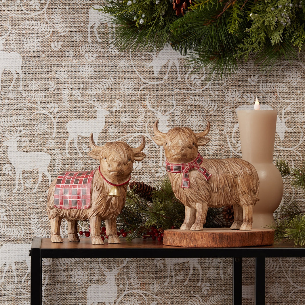 Two Highland Cow figurines with plaid scarf and blanket accents, designed for holiday and winter decor, placed on a table with festive decorations in the background.