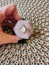 Load image into Gallery viewer, Hexagonal pink jewelry box with a ring featuring a central opal gemstone held by a hand on a textured surface.

