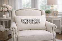 Load image into Gallery viewer, Beige armchair with a pillow displaying 'Doylestown' and coordinates in a home setting.
